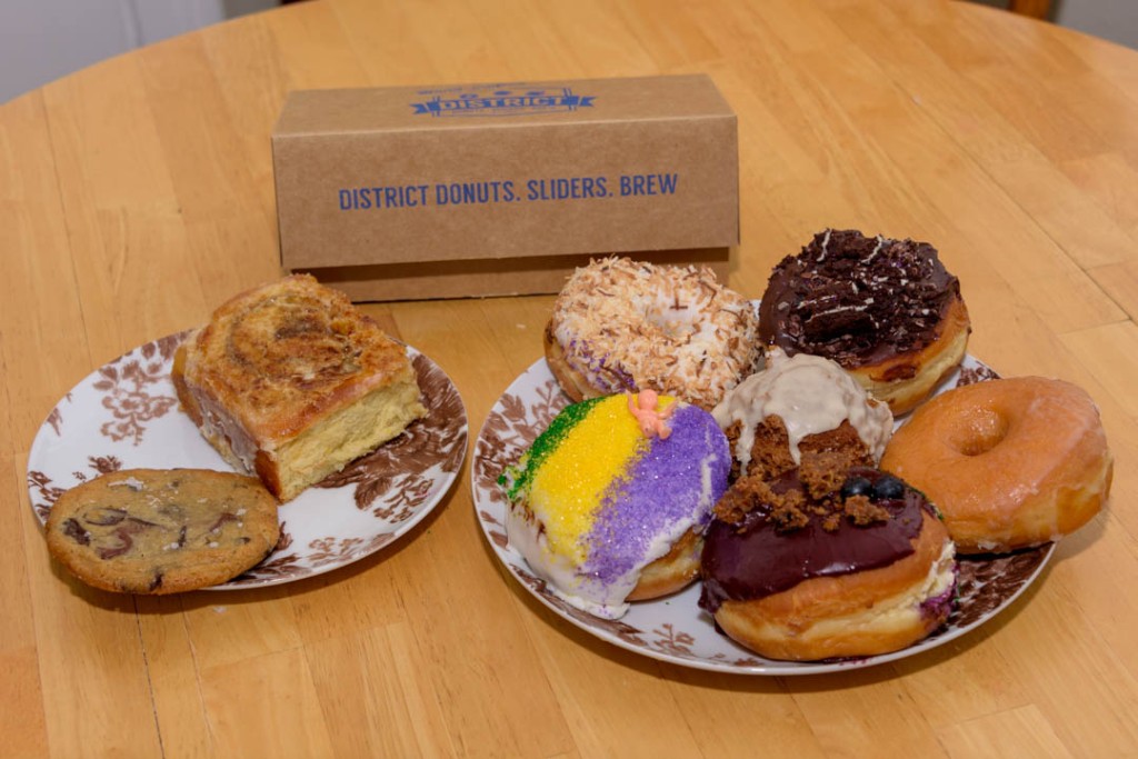 Two plates of desserts on a table, including donuts, cinnamon roll and cookie, with District Donuts, Sliders, Brew box in the background.